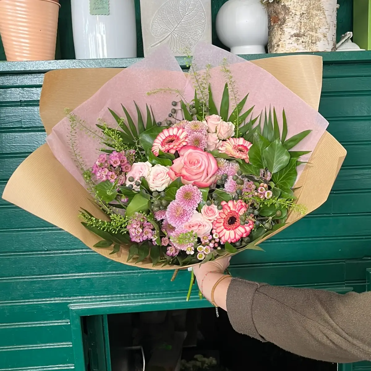 Bouquet rose emballé de papier tenu devant la devanture verte d'une boutique de fleuriste