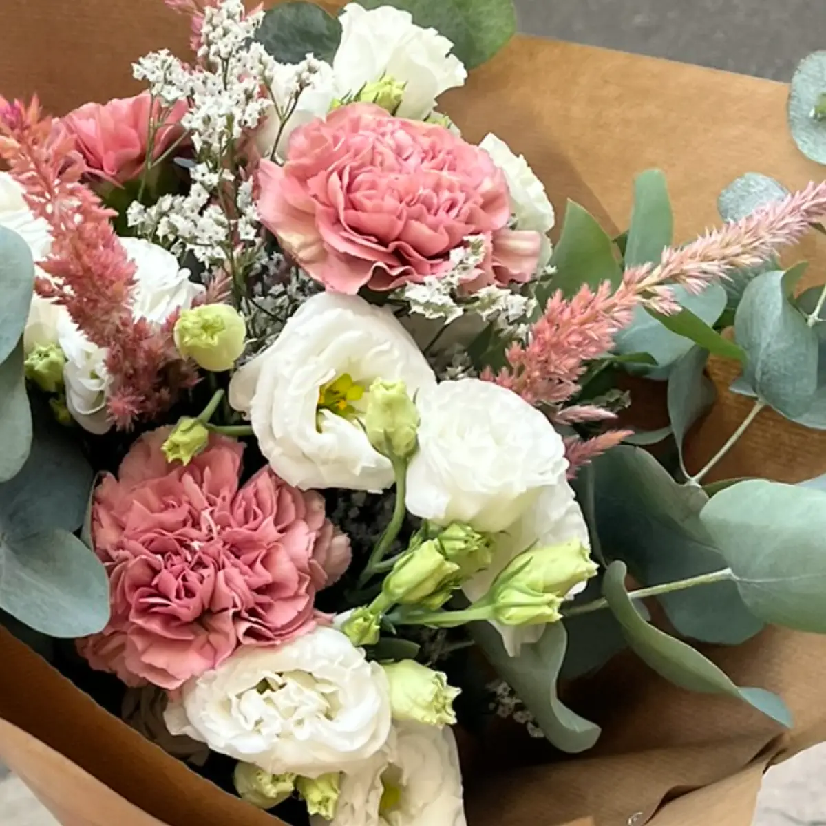 Bouquet en papier kraft où des lisianthus blancs voisinent avec des œillets roses, de la célosie et de l'eucalyptus