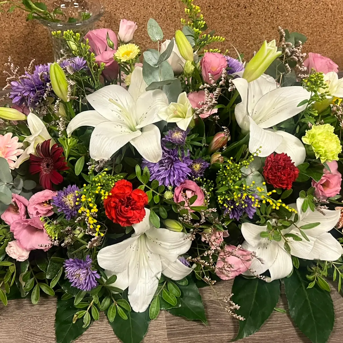 Raquette de deuil ovale posée sur un établi, où une dizaine de lys blancs ouverts dominent des asters violets, des lisianthus roses et des œillets rouges