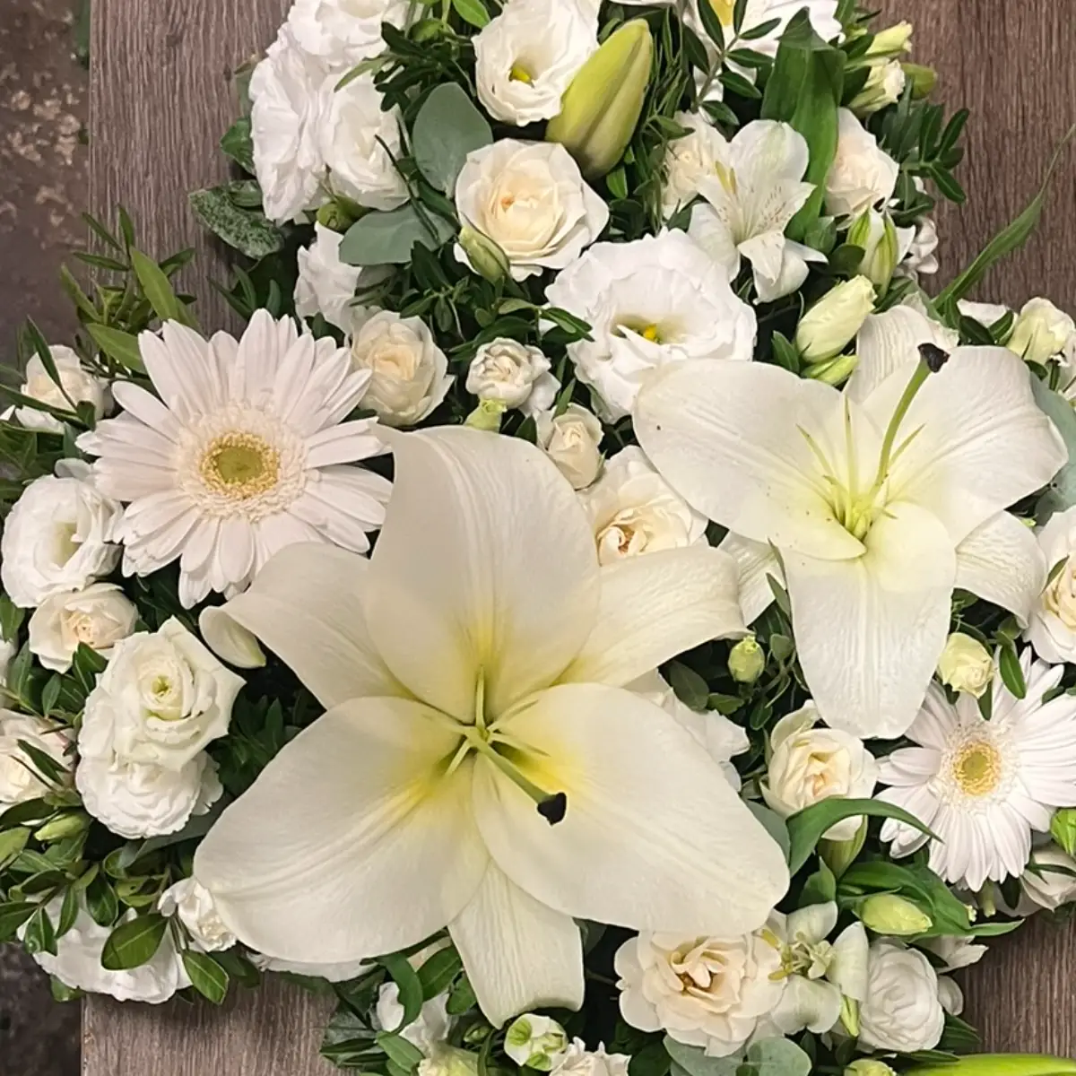 Croix de deuil fleurie de lys blancs, roses branchues, gerbera et lisianthus, posée sur un plan de travail en bois
